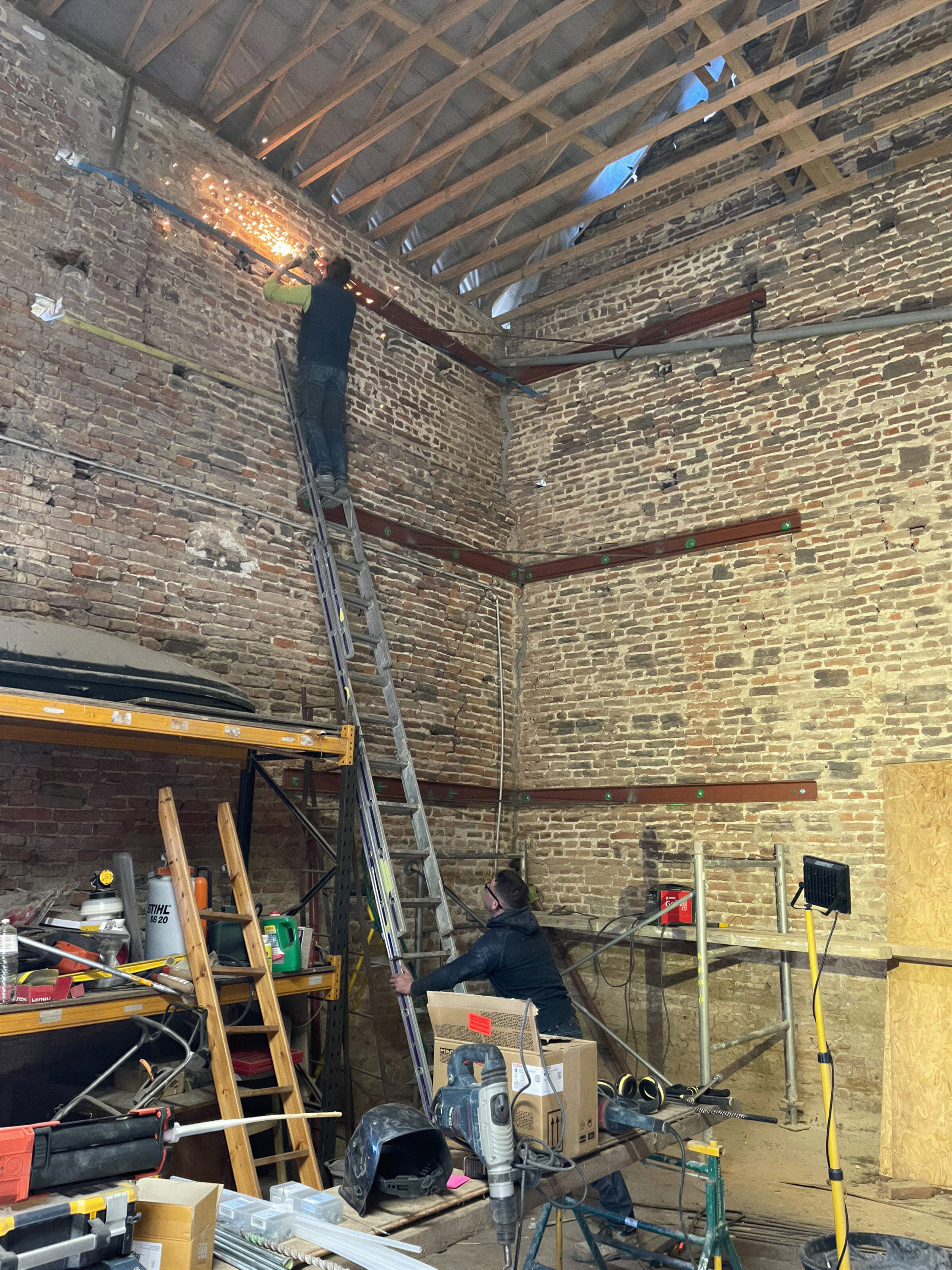 Artisans au travail sur un mur en briques et moellons, sous une charpente en rénovation