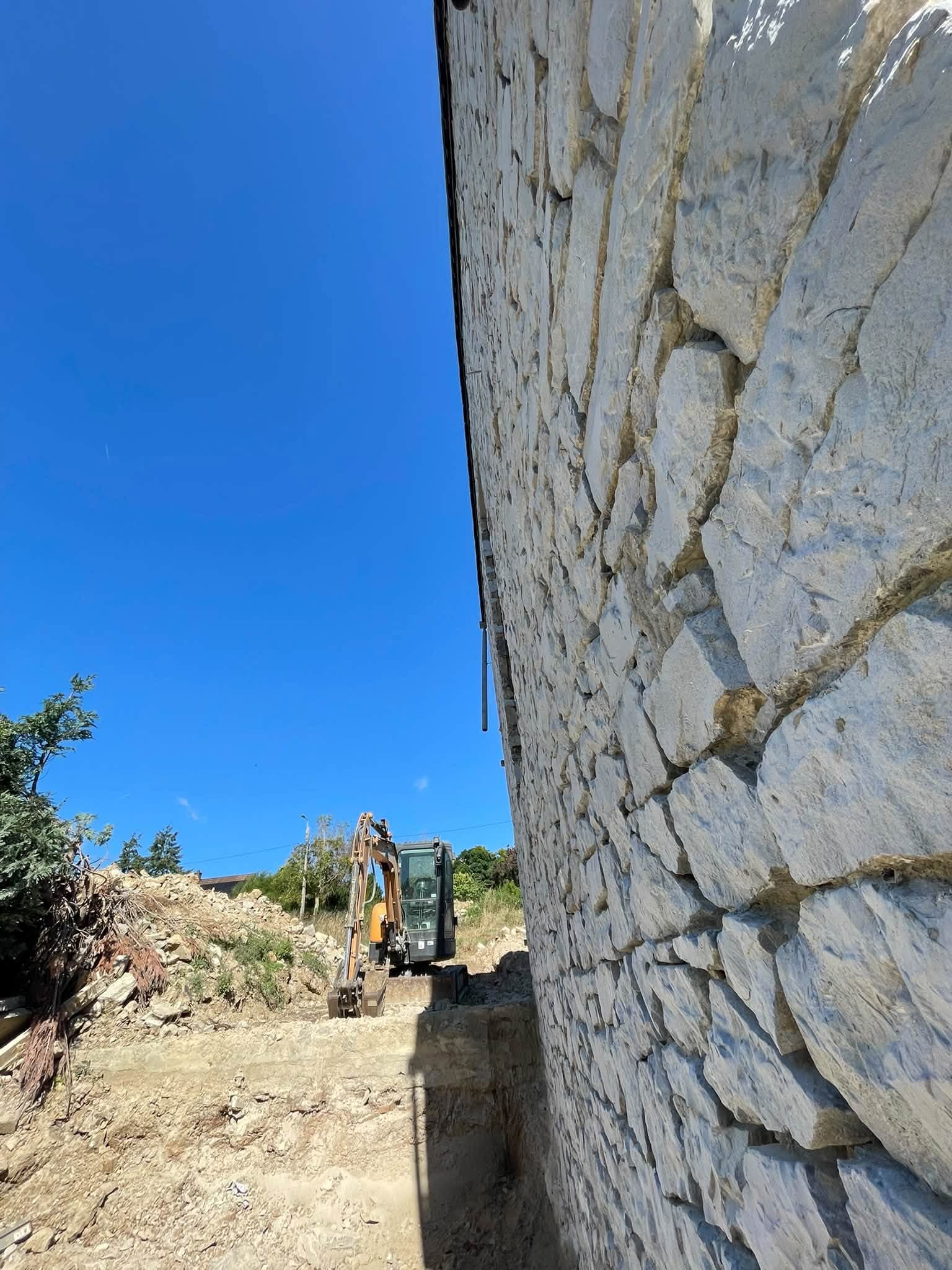 Haut mur en pierre restauré, vu en contre-plongée, avec une pelleteuse au pied